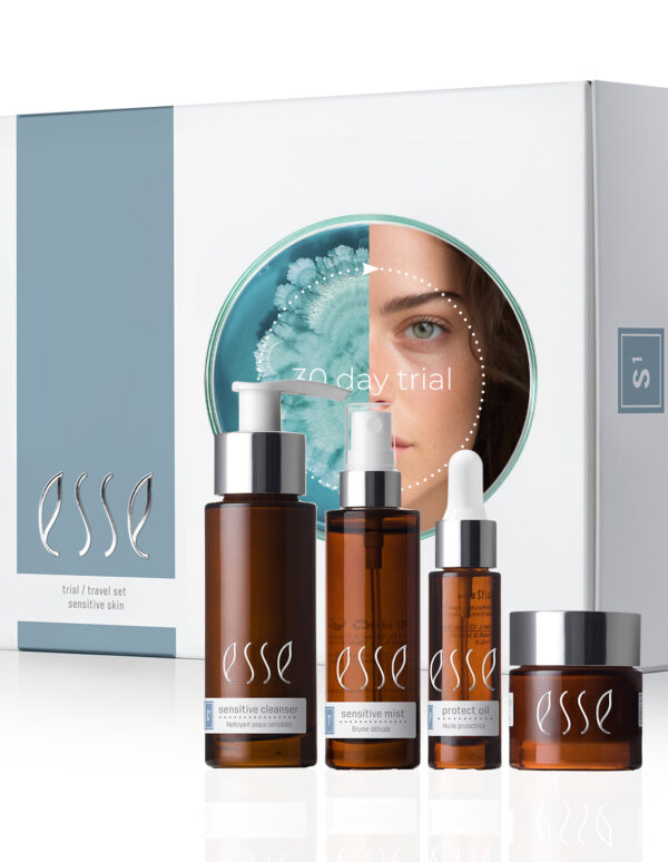 The Esse Sensitive Skin Trial / Travel Set is displayed, featuring four brown bottles and jars in various sizes positioned in front of white and blue packaging. The packaging highlights an image of a woman’s face partially obscured by a flower.