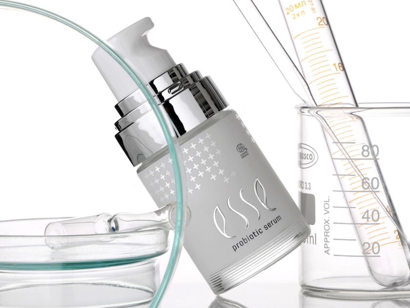 A bottle of Esse probiotic serum is displayed among laboratory glassware, highlighting a scientific approach to skincare and addressing the causes of accelerated ageing for healthier, youthful-looking skin.