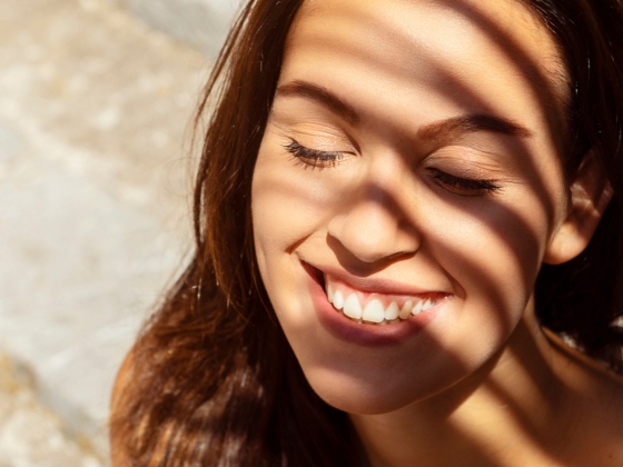 A woman with long brown hair smiles with her eyes closed as sunlight and shadows from blinds fall across her face, radiating the natural glow of live probiotic skincare.