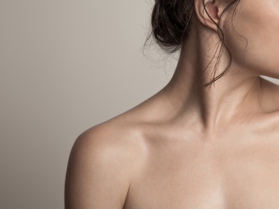 A close-up of a woman's bare shoulder, neck, and lower face against a neutral background highlights the natural beauty of her skin microbiome, with her hair partially tied back and a loose curl visible.